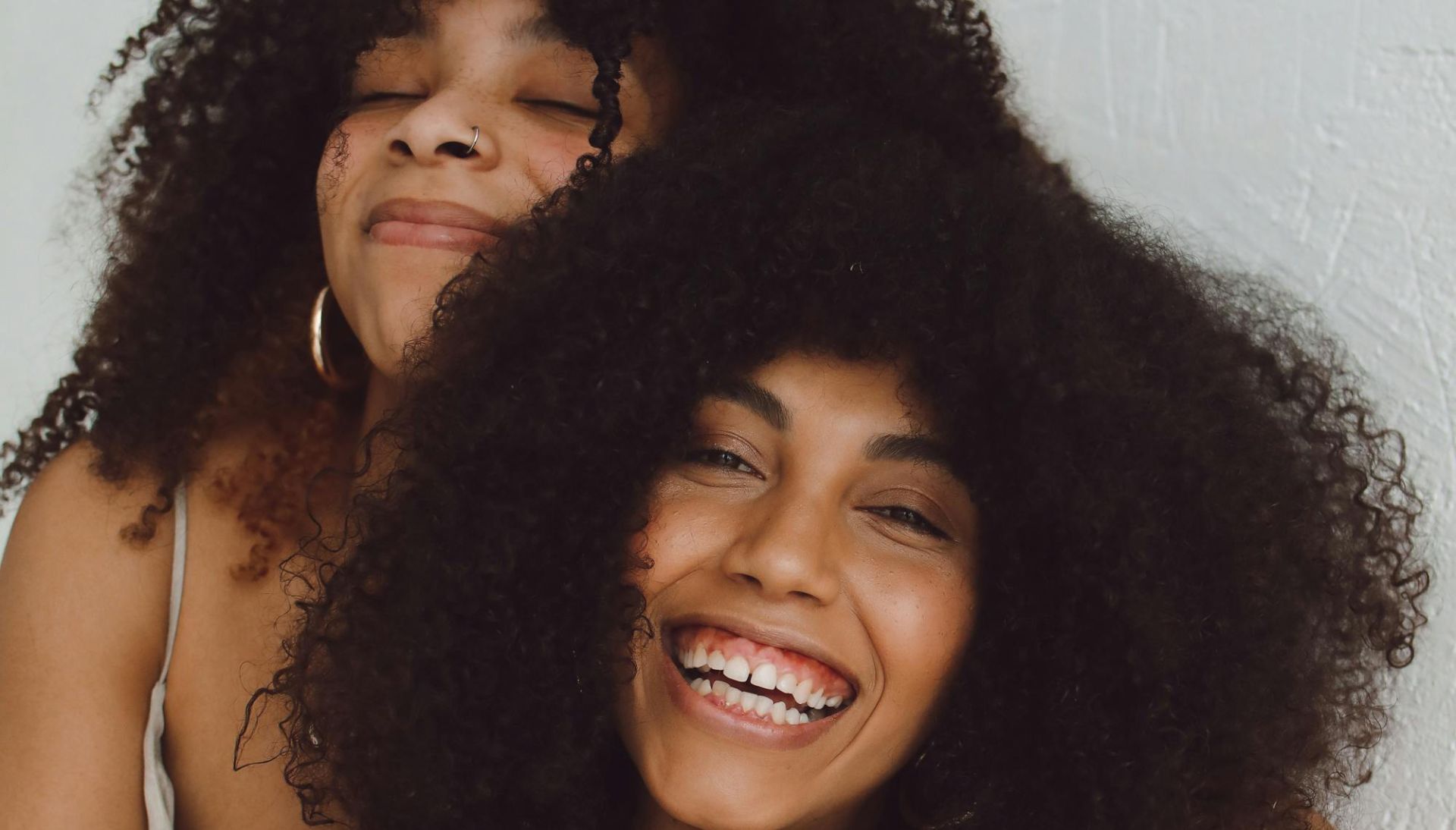 Two smiling women with curly hair embrace against a white wall, radiating joy and connection.