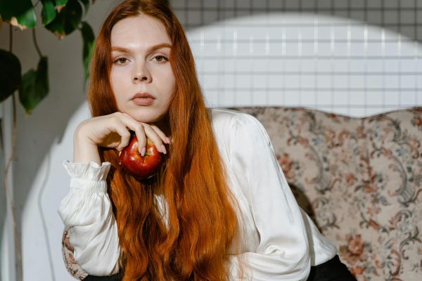 Elegant redhead woman holding an apple, posing in soft indoor lighting.