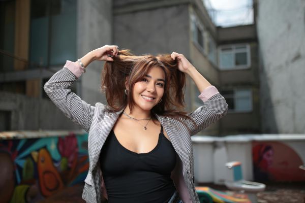A joyful woman with a checkerboard jacket smiles while holding her hair outdoors in a vibrant urban setting.